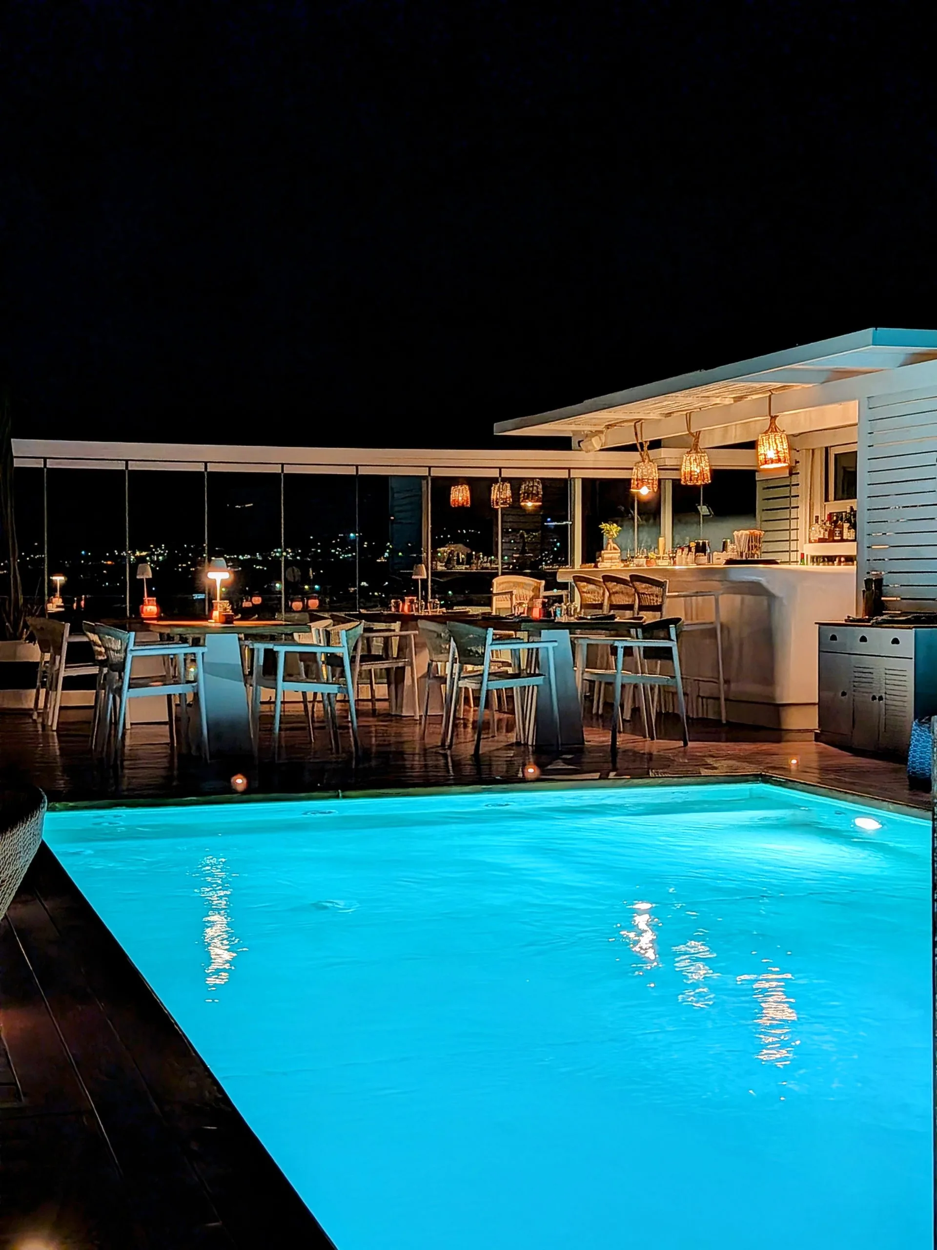 Elegant rooftop lounge at night featuring a luminous turquoise pool, chic bar seating, warm pendant lighting, and panoramic skyline views.